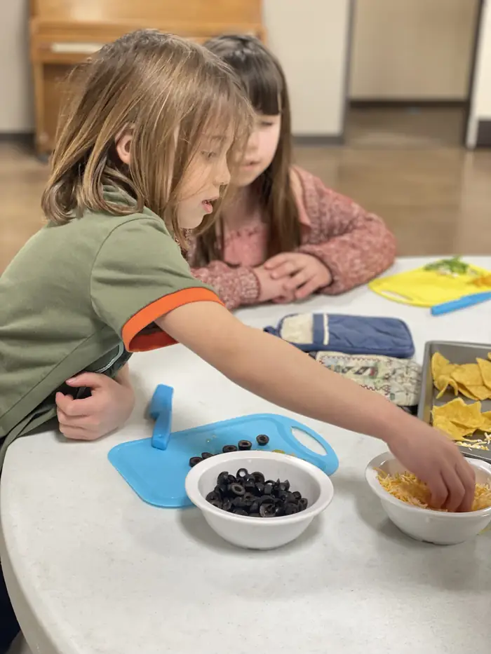 Two Cornerstone students adding cheese and olives to a tray of nachos in a cooking lesson