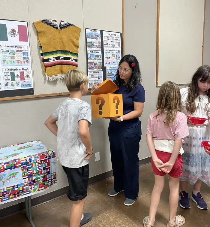 A Missionaries in Training teacher opening a mystery box for a student beside a display about Mexico